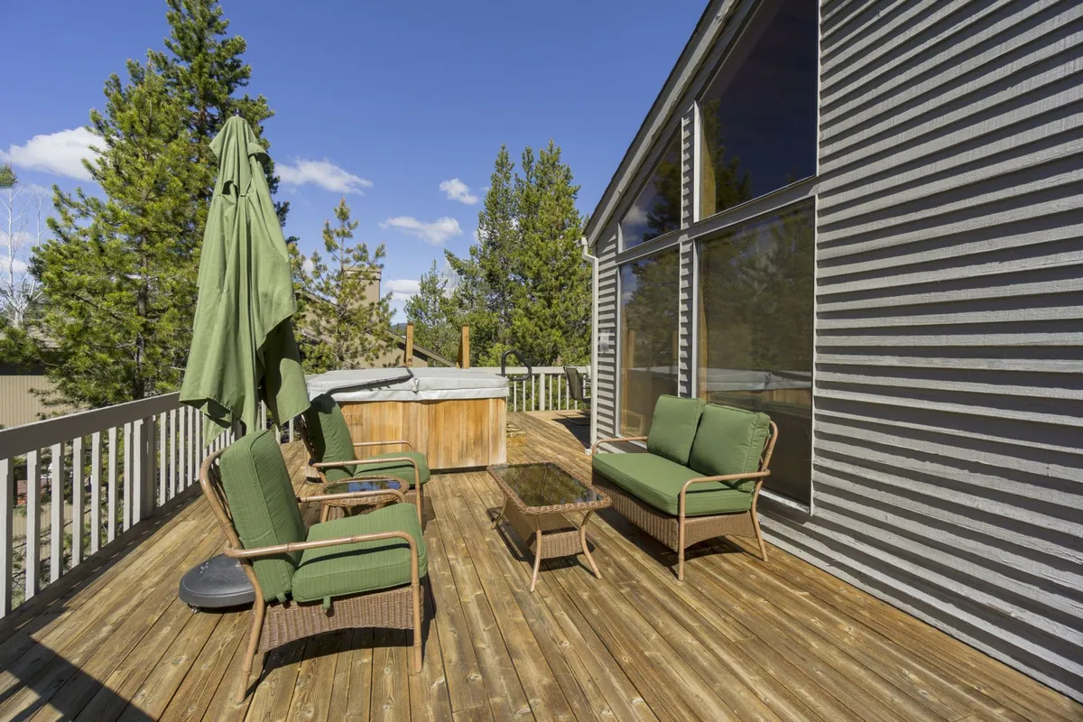 An expansive wooden deck featuring comfortable seating, a shaded umbrella, and a hot tub, all nestled amidst greenery. The scene suggests a tranquil space for relaxing after a day of exploring, with plenty of room for both people and pets to lounge outdoors.