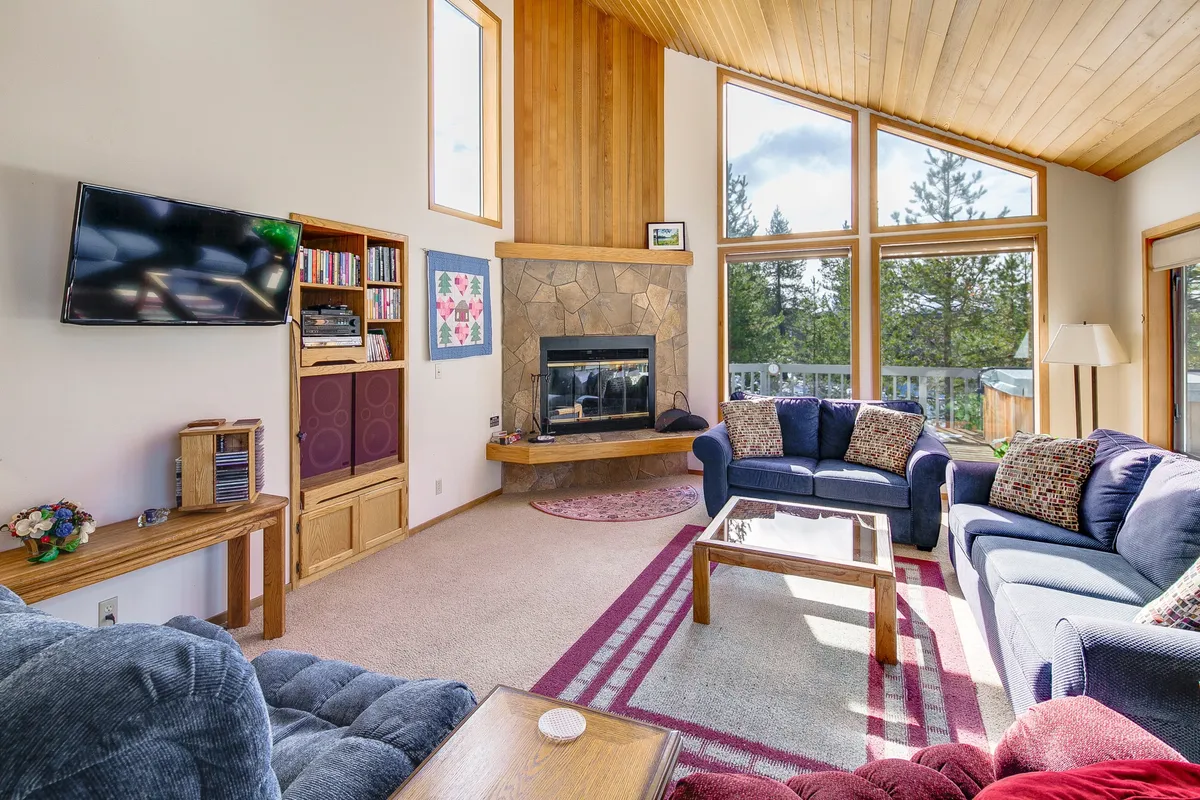The image reveals a cozy living room with vaulted, wood-paneled ceilings, a crackling fireplace as the centerpiece, plush sofa seating, a welcoming coffee table, and expansive windows that invite in natural light and views of the outdoors. The scene exudes warmth and a sense of togetherness, perfect for relaxing, gathering, or simply watching the sunset after a day of adventure.