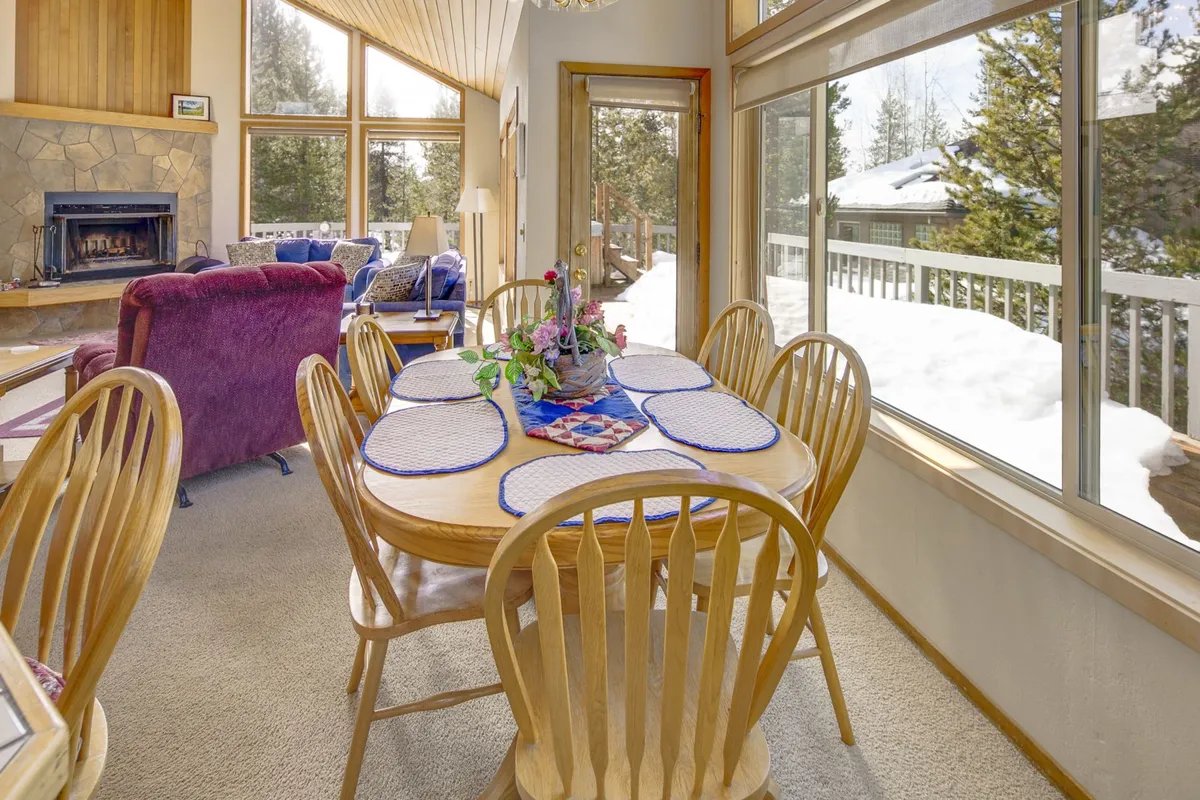 A view of the dining area and living room, featuring a dining table, a nearby fireplace, a sofa, and expansive windows. The setting exudes warmth and is ideal for family meals, board games, or reading by the fire with snowy or scenic views visible beyond.