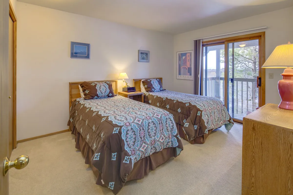 A cozy twin bedroom featuring two twin beds, a lamp between them, a sliding glass door, nightstands, and decorative wall art. The setting is comfortable and designed for both relaxation and socializing, with light streaming through the door.