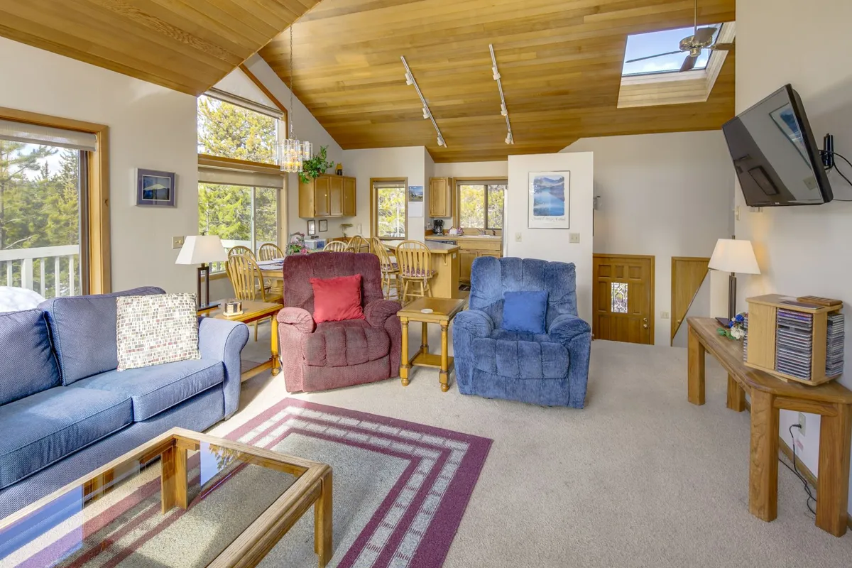 An open-concept living area showcasing a plush sofa, recliners, a coffee table, and an adjacent kitchen area visible beyond. The space is accented by windows letting in daylight, reinforcing a seamless flow between living and kitchen spaces ideal for gatherings.