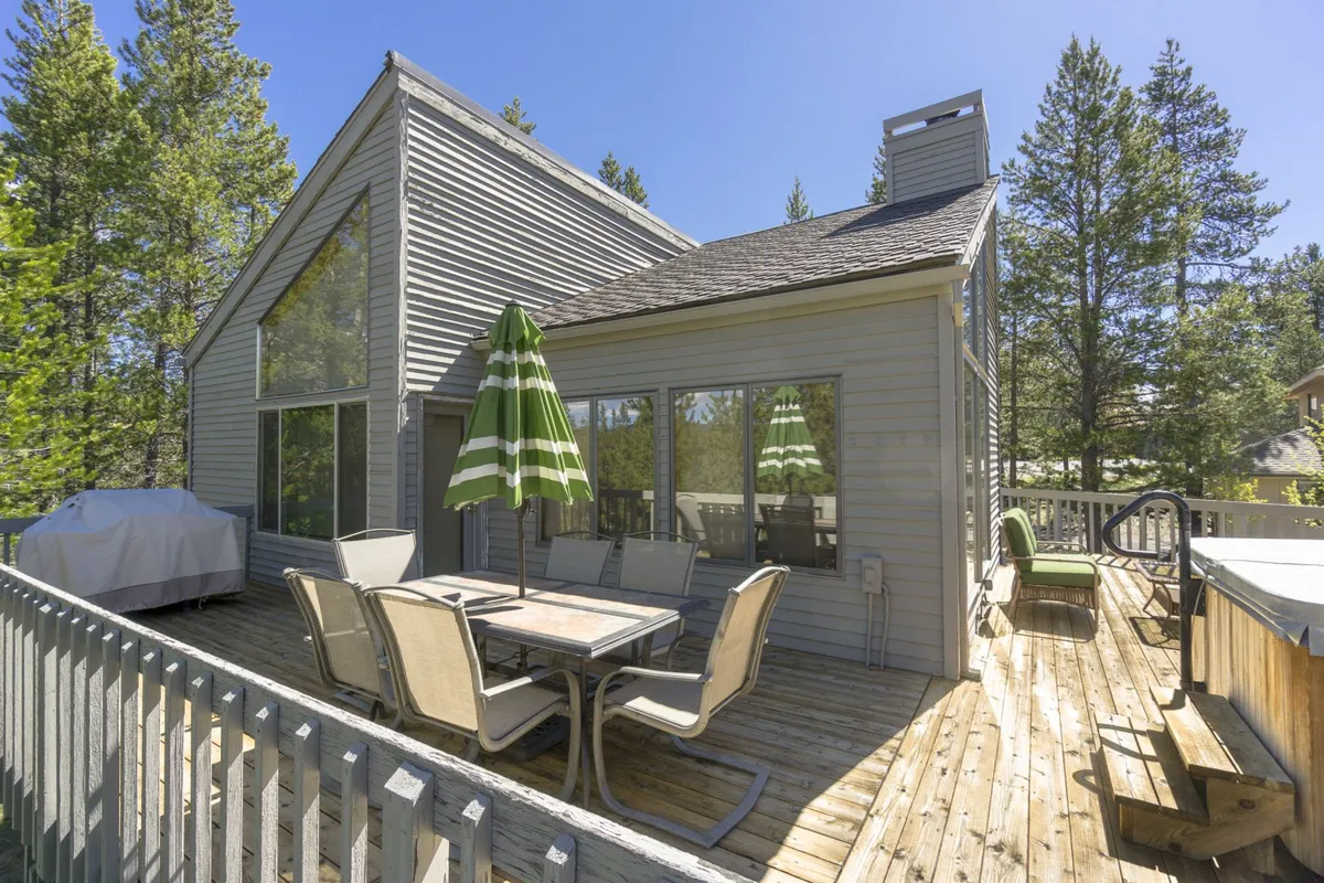 An inviting deck scene featuring an outdoor dining table with a striped umbrella, a hot tub, and surrounding trees. The image communicates a lifestyle of relaxation, outdoor dining, and group gatherings amidst natural beauty.