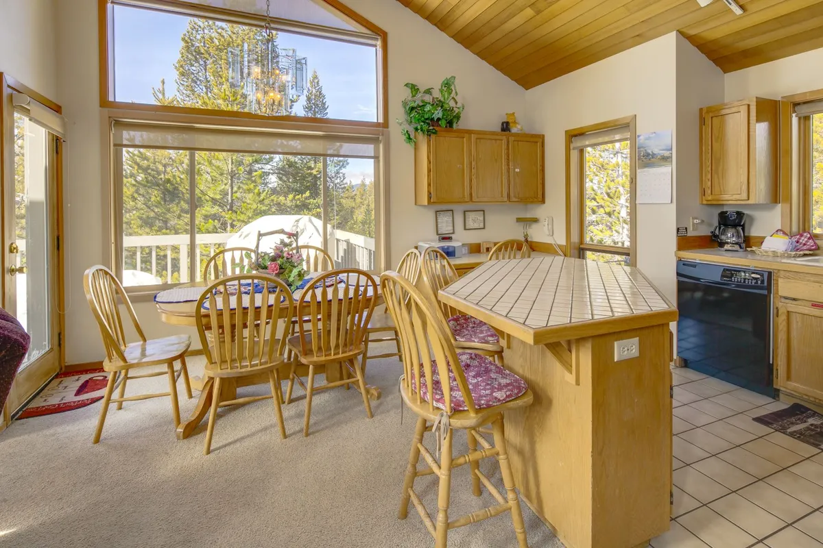 A spacious, sunlit open-plan kitchen and dining area. The image shows a large dining table set beside expansive windows that offer outdoor views. The adjacent kitchen counter seamlessly connects to the dining area, creating a welcoming space for group meal prep and shared family meals.
