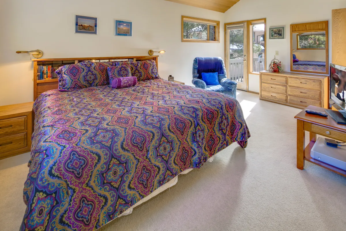 This image features a large bed with colorful bedding, a wooden dresser, a comfortable blue chair, and expansive windows filling the room with natural light. The space is warm, inviting, and suggests a restful master bedroom retreat.